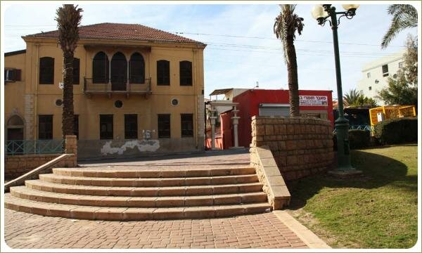 Лестница из светлого камня в форме полукруга и зелёная лужайка возле каменый стены и дом жёлтого цвета