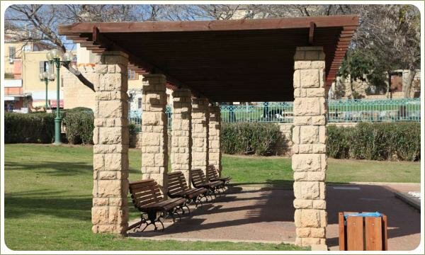 Лёгкое строение на каменных столбах с деревянным крышей и скамейки на фоне площадки и лужайки