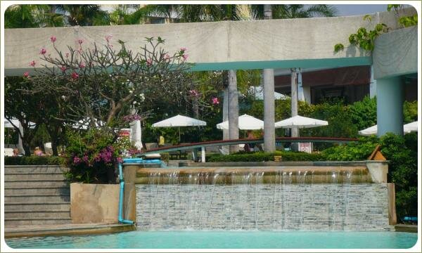 Водный каскад для бассейна. Сад в Таиланде Падающая вода на фоне стены у бассейна и деревья и зонты от солнца на берегу