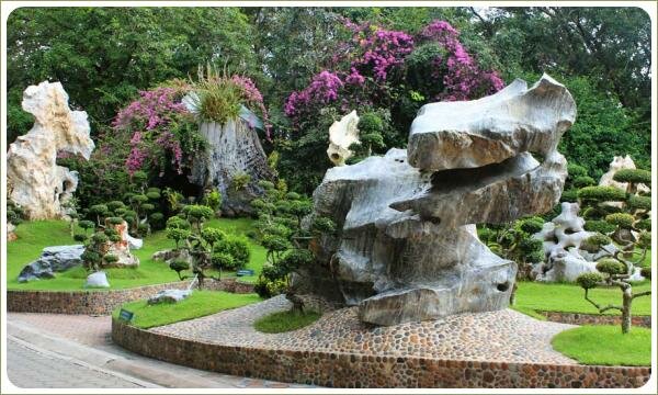 Камень в виде сказочного животного серого цвета на фоне пышных растений сиреневого цвета на площадке парка