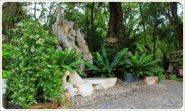 Скамья из светлого камня и камень в форме спинки трона в зарослях тропических растений на фоне мощёной площадки