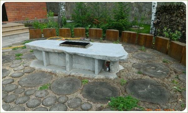 Сооружение из светлого камня с металлической вставкой на фоне площадки мощёной спилами