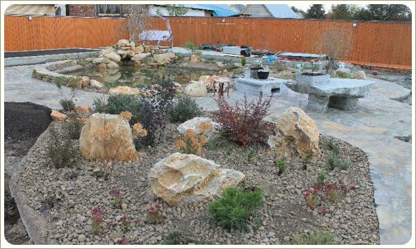 Крупные камни светлого цвета на гравийной отсыпке среди растений и площадка с каменными сооружениями