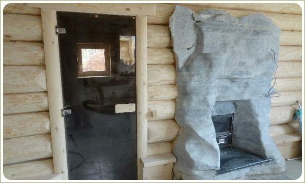 На стене из светлого дерева смонтирована из серого камня печка