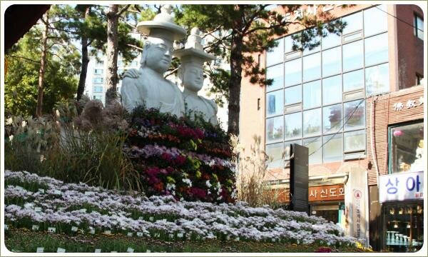 Скульптуры из светлого материала с постаментом из разноцветных цветов на поляне с сиреневыми и розовыми и зелёными растениями 