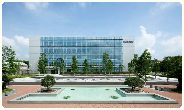 Прямоугольный водоём с маленькими растениями на фоне площади с разного цвета покрытием и штамбовых деревьев на фоне остеклённого здания