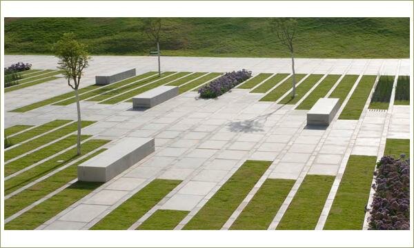 Скамейки похожие на простые прямоугольные короба на фоне газонов в длинных грядках и площади мощёной прямоугольными плитами