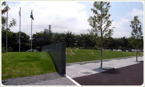 Облицованная блестящим камнем стена на фоне холма-газона и гравия и тротуарной плитки и коричневого покрытия и серого покрытия и деревьев