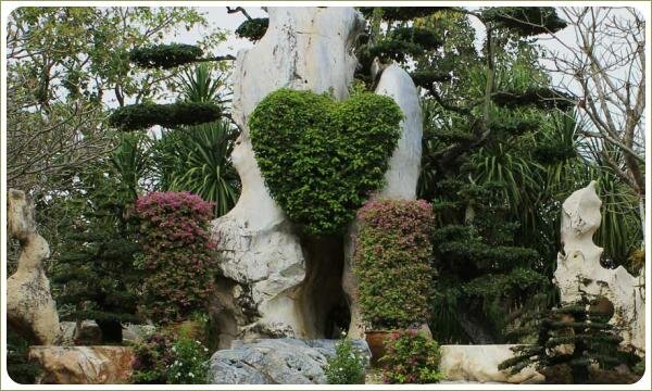 Стриженный в в форме сердца кустарник на фоне причудливого светлого камня и розовоцветущих кустарников и плоских камней и пальмв