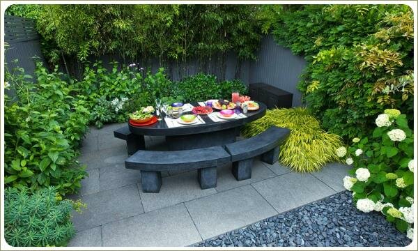 На площадке мощёной серым камнем набор садовой мебели темно серого цвета на фоне миксбордеров с растеними различных оттенков зелени