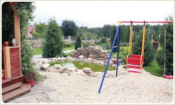 Детская площадка на фоне каменистой отсыпки и водоёма с горкой и хвойных расений и беседки