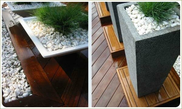 Серые горшки с крупными ветвистыми растениями на деревянных постаментах на мощёной камнем площадке на фоне стены строения