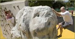 Свелая скульптура из композитного матерала похожая на каменную глыбу с углублениями для лазания на фоне стены с приспоблением для лазания на площадке с жёлтым покрытием