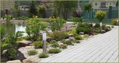 Каменистый садик с растениями и светлым гравием на фоне большого пруда